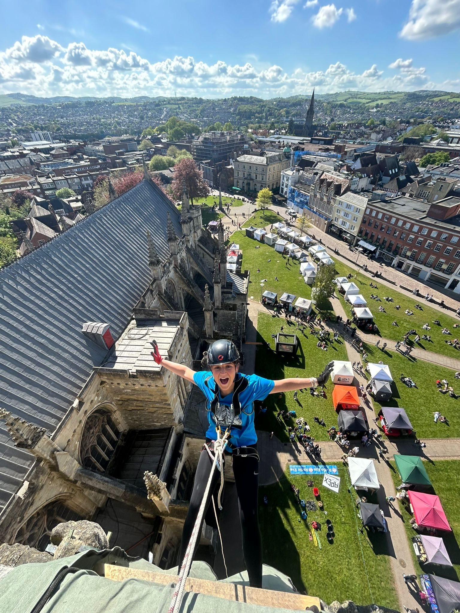 Charity abseiler with arms outstretched high on a cathedral tower above the event village on the green below
